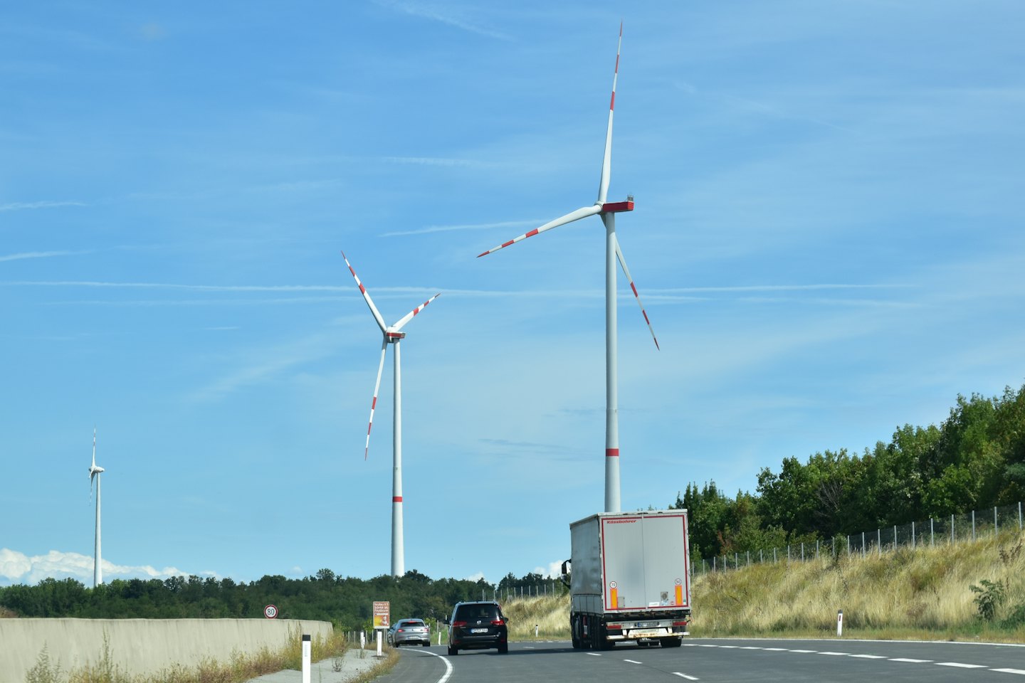 Wind turbines stand near a highway with vehicles driving past.